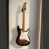 Elektrická kytara s tłem v barvě sunburst a bílým pickguardem. Na hlavě hředu je viditelné logo Fender. Nástroj má javorový hmatník a tři snímače typ…