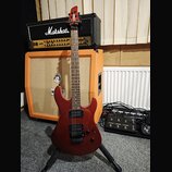 Tmavě červená elektrická kytara s hnědým hmatníkem a černými snímači. Na hlavě je bílý nápis Ibanez. V pozadí je kytarový zesilovač Marshall s nápise…