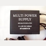 Černý hranatý zdroj napájení s textem MULTI POWER SUPPLY, SEPARATE ISOLATED a 6 x 10V DC/100mA. Dole je uveden nápis STARBUCK. Povrch zařízení má jem…