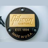 Kulatý černý kovový kryt s nápisy v zlaté barvě. Uprostřed je zlatý obdélníkový štít s vyrytým logem Gibson a nápisem CUSTOM. Pod ním jsou vloženy te…