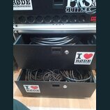 Černý rackový kufřík se dvěma vysunutými šuplíky, které obsahují svitek černých kabelů. Na přední straně šuplíků jsou nalepené bílé samolepky s nápis…