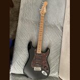 Černá elektrická kytara s hnědým hnedým hmatníkem a tmavě hnědým vzorovaným pickguardem. Na hlavě je viditelné logo "Squier". Kytara má tři snímače,…