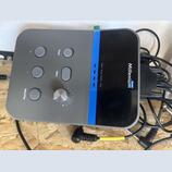 Šedý elektronický modul s černým displejem a modrým svislým pruhem. Na zařízení je bílý nápis Millenium a modrý štítek s nápisem Drums. Ovládací pane…