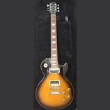 Elektrická kytara typu Les Paul s barevným přechodem od žluté přes oranžovou k černé (sunburst). Na hlavě hmatníku je bílý nápis Gibson a pod ním pod…