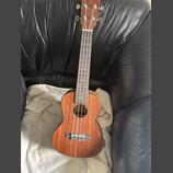 Akustické ukulele s hnědým dřevěným povrchem a čtyřmi strunami. Nástroj má hnědý krk s bílými tečkovými značkami na hmatníku a stříbrné ladičky. Na s…