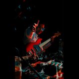 Fotografie zachycuje muže s červenou elektrickou basovou kytarou. Nástroj má červené tělo, bílý pickguard a světle hnědý hmatník. Na snímku nejsou vi…
