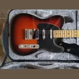 Elektrická kytara typu Telecaster v barevném přechodu od červené přes oranžovou k černé s černým pickguardem a javorovým hmatníkem. Nástroj je umístě…