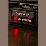 Černý kytarový kombozesilovač značky Blackstar z řady HT MkII. Na předním panelu jsou bílé a zelené ovratové knofíky s popisky POWER, REVERB LEVEL, E…