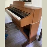 Digitální piano v dřevěném korpusu s hnědým povrchem. Nástroj má bílé a černé klávesy a tři pedály v dolní části. Na povrchu nejsou vidět žádné značk…