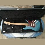 Modrá elektrická kytara typu Stratocaster s červeným trochlodňovým pickguardem v černém tvrdém kufru. Na hlavě kytary je nápis Fender STRATOCASTER. N…
