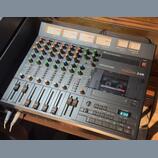Šedý vícetrackový magnetofon s integrovaným mixážním pultem. Na zařízení jsou nápisy PORTASTUDIO a TASCAM 246, v kazetovém mechanismu je vložena kaze…