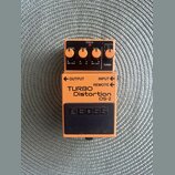 Kytarový efekt pedál oranžové barvy s černým spodním panelem. Na zařízení jsou nápisy TURBO Distortion DS-2 a logo BOSS. Horní část obsahuje tři ovlá…