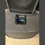 Detail basového reproduktoru v šedém čalounění z látky. Na horní části je černá plastová rukojeť s modro-bílou nálepkou s nápisem Megaton. V horní a…