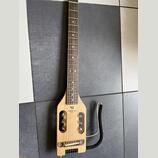 Světle hnědá elektrická kytara s krátkým tělem a dlouhým hnedým hmatníkem. Na těle je černý nápis TRAVELER GUITAR a nad ním logo ve tvaru trojúhelník…