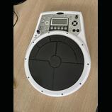 Bílý elektronický perkusní pad Roland HandSonic 10 s velkým černým gumovým playing polem. V horní části je ovládací panel s displejem, několika tlačí…