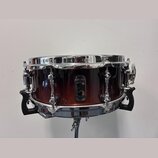 Kytarový snare drum s korpusem v červenohnědém přechodovém odstínu a chromovanými obručemi. Uprostřed korpusu je černý štít s bílým logem a nápisy BL…