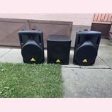 Tři černé reproduktorové bedny z plastu umístěné vedle sebe na betonovém povrchu. Uprostřed stojí menší hranatý subwoofer a po stranách dva větší top…