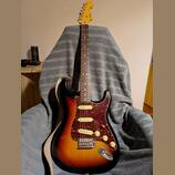 Elektrická kytara s trupem v barevnosti sunburst a hnědým hmatníkem. Kytara má červeně mramorovaný pickguard a na hlavě je viditelné logo Fender. Pov…