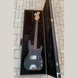 Černá elektrická basová kytara v černém tvrdém kufru. Na hlavě hmatníku je nápis Fender. Nástroj má tmavý hmatník, bílý pickguard a chromované hardwa…