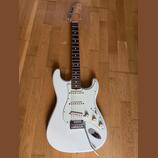 Bílá elektrická kytara typu Stratocaster s bílým pickguardem a hnedým hmatníkem. Na hlavě je vidět nápis Fender. Nástroj má tři snímače, tři ovládací…