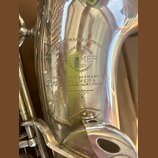 Detail stříbrného povrchu saxofonu s vyrytou značkou. Uprostřed je logo s nápisy HENRI SELMER PARIS obklopené věncem a nadepsáno BREVETÉ FRANCE & ETR…