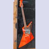 Oranžová elektrická kytara s hranatým tělem v podobě šipky, položená v černém kufru. Na hlavě hředu je zlaté logo Gibson. Kytara má dva humbuckery, z…