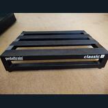 Černý kovový pedalboard s mřížkovou konstrukcí a vložkami z černého textilního popruhu. Na přední hraně je bílý nápis pedaltrain® a pod ním menší tex…
