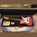 Červená elektrická kytara s bílým pickguardem v černém polstrovaném kufru. Na hlavě hmatníku je nápis Fender Stratocaster. V kufru vedle nástroje lež…