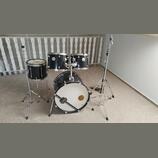 Černá bicí souprava s chromovaným kováním, která obsahuje basový buben, dva tom-tomy a jeden snare drum. Na přední straně basového bubnu je bílý kože…
