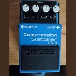 Modrý efekt pedál s nápisem Compression Sustainer CS-3. V horní části jsou čtyři bílé ovládací knoflíky, červená kontrolka a nápisy CHECK, AT, SUS, M…