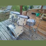 Bílá bicí souprava s chromovaným kováním, sestávající z basového bubnu, dvou tomů a malého bubnu. Na činelech je vidět nápis MEINL a LIVE SOUND. Nást…