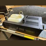 Černý mlhový generátor s bílou plastovou nádrží na tekutinu. Na boku přístroje je bílý nápis F-100 PERFORMANCE FOG GENERATOR a dvě varovné žluté znač…