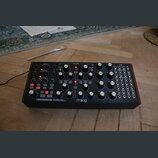 Černý analogový polyrytmický syntetizátor s nápisy SUBHARMONICON a moog. Přístroj obsahuje řadu bílých otočných ovladačů, červených podsvícených tlač…