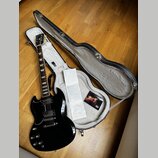 Černá elektrická kytara s typickým tvarem SG, equipped s dvěma humbuckery a čtyřmi ovládacími knoflíky. Na hlavě hmatníku je vidět nápis Gibson. Kyta…