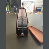 Hnědý mechanický metronom s bílým úzkým úželem a černým spodním panelem. Na přístroji jsou nápisy "Wittner METRONOM", "taktell PICCOLO" a označení te…