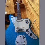 Modře třpytivá elektrická kytara s bílým pickguardem v hnědém polstrovaném kufru. Na kovovém krytu spodního tremola je nápis Fender. Nástroj má hnědý…
