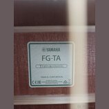 Detail štítku uvnitř akustické kytary. Na bílém pozadí s zeleným rámem je uveden nápis YAMAHA, model FG-TA a označení TransAcoustic v oválu. Dole je…