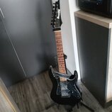 Černá elektrická kytara s hnědým hmatníkem umístěná na stojanu. Na hlavě je bílý nápis Ibanez. Kytara má tři snímače, černý pickguard a připevněný še…