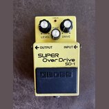 Žlutý kytarový efekt pedál značky BOSS, model SUPER Over Drive SD-1. Na horní části jsou tři černé ovládací knoflíky s bílými vršky a nápisy LEVEL, T…
