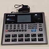 Stříbrný bicí automat značky Alesis, model SR18. Na předním panelu je nápis HIGH DEFINITION STEREO MULTI-SAMPLED DRUM MACHINE WITH DYNAMIC ARTICULATI…