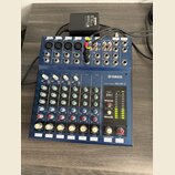 Modrý mixovací pult s nápisem YAMAHA MIXING CONSOLE MG10/2. Horní panel obsahuje různé vstupy a výstupy, včetně konektorů XLR a jack, s popisky jako…