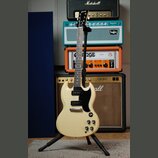 Elektrická kytara s bílým trupem a černým pickguardem umístěná na černém stojanu. Na hlavě hmatníku je viditelné logo Gibson. V pozadí jsou čtyři kom…