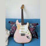 Růžová elektrická kytara typu Stratocaster s bílým pickguardem a hnědým hmatníkem. Povrch instrumentu vykazuje výrazné opotřebení, zejména v podobě o…