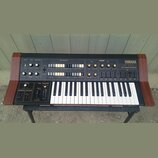 Černý analogový syntetizátor s dřevěnými bočními panely a bílo-černou klaviaturou. Na pravé straně je nápis YAMAHA CS15D DUAL CHANNEL SYNTHESIZER. Př…