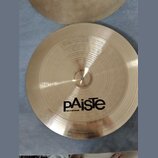 Zlatý kovový činel s koncentrickým žebrováním povrchu. Na spodní části je černý nápis pAisTe a pod ním menší text MADE IN SWITZERLAND. Povrch je v do…