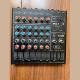 Černý mixážní pult s šesti kanály. Na pravé straně je bílý nápis MMR 13 model V a logo SEAK. Zařízení obsahuje řadu barevných otočných ovladačů v ora…