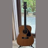 Akustická kytara s přirozenou dřevěnou barvou korpusu a černým hmatníkem. Na hlavě je bílý nápis YAMAHA a dekorativní ornament. Kytara má černý trsac…