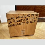 Dřevěný nástroj ve tvaru kostky s přirozenou barvou dřeva. Na přední straně je černý nápis SCHLAGWERK PERC 26"BOOSTER BOXX GERMANY2 IN ONE O a ve spo…