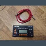 Černý digitální ladič a metronom značky BOSS, model TU-80. Zařízení má LCD displej, oranžová tlačítka a nápisy POWER, TUNER, METRONOME, SOUND SELECT,…