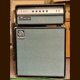 Basový kombozesilovač značky Ampeg skládající se z hlavy a boxu. Na předním panelu hlavy jsou ovládací prvky, konektory a nápis Ampeg. Box je potažen…